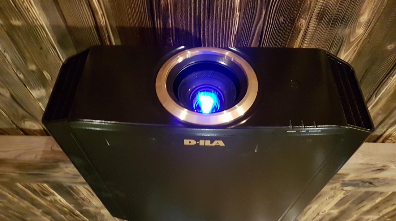 Czarny projektor D-ILA zamontowany na drewnianym suficie z widocznym obiektywem z niebieskim światłem.