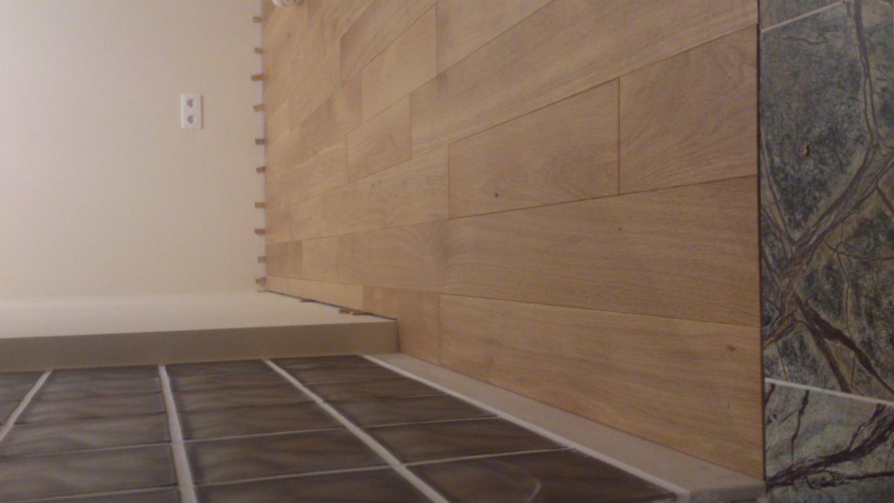 Układanie drewnopodobnych paneli ściennych w łazience, widoczne płytki podłogowe i fragment ściany wykończonej zielonym marmurem