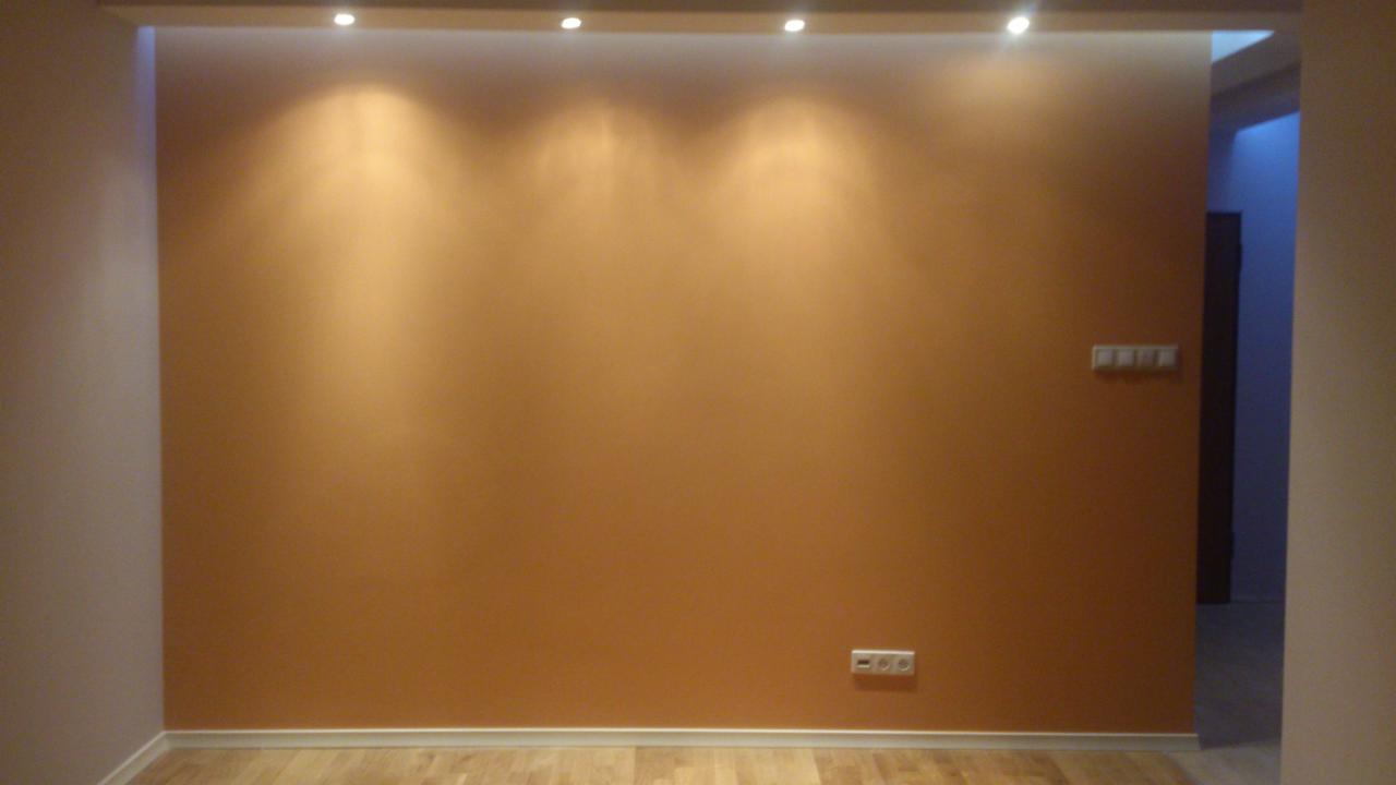 Pomarańczowa ściana w pokoju z drewnianą podłogą, oświetlona przez punktowe światła w suficie, widoczne gniazdka elektryczne i włączniki.