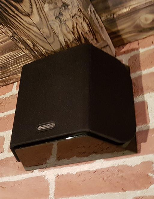 Czarny głośnik Monitor Audio zamontowany na ceglanej ścianie pod drewnianą belką stropową.