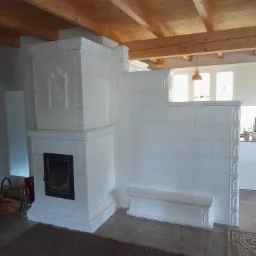 Biały, kaflowy piec z paleniskiem i ozdobną płaskorzeźbą, zintegrowany z niską ścianką działową i siedziskiem, widoczne drewniane belki stropowe