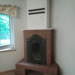 Dariusz Majak usługi zduńskie - Murowany kominek z cegły z czarnymi żeliwnymi drzwiczkami, zwieńczony białym prostokątnym obudowaniem z poziomymi szczelinami wentylacyjnymi, umieszczony w rogu pokoju obok okna z kwiecistą zasłonką.