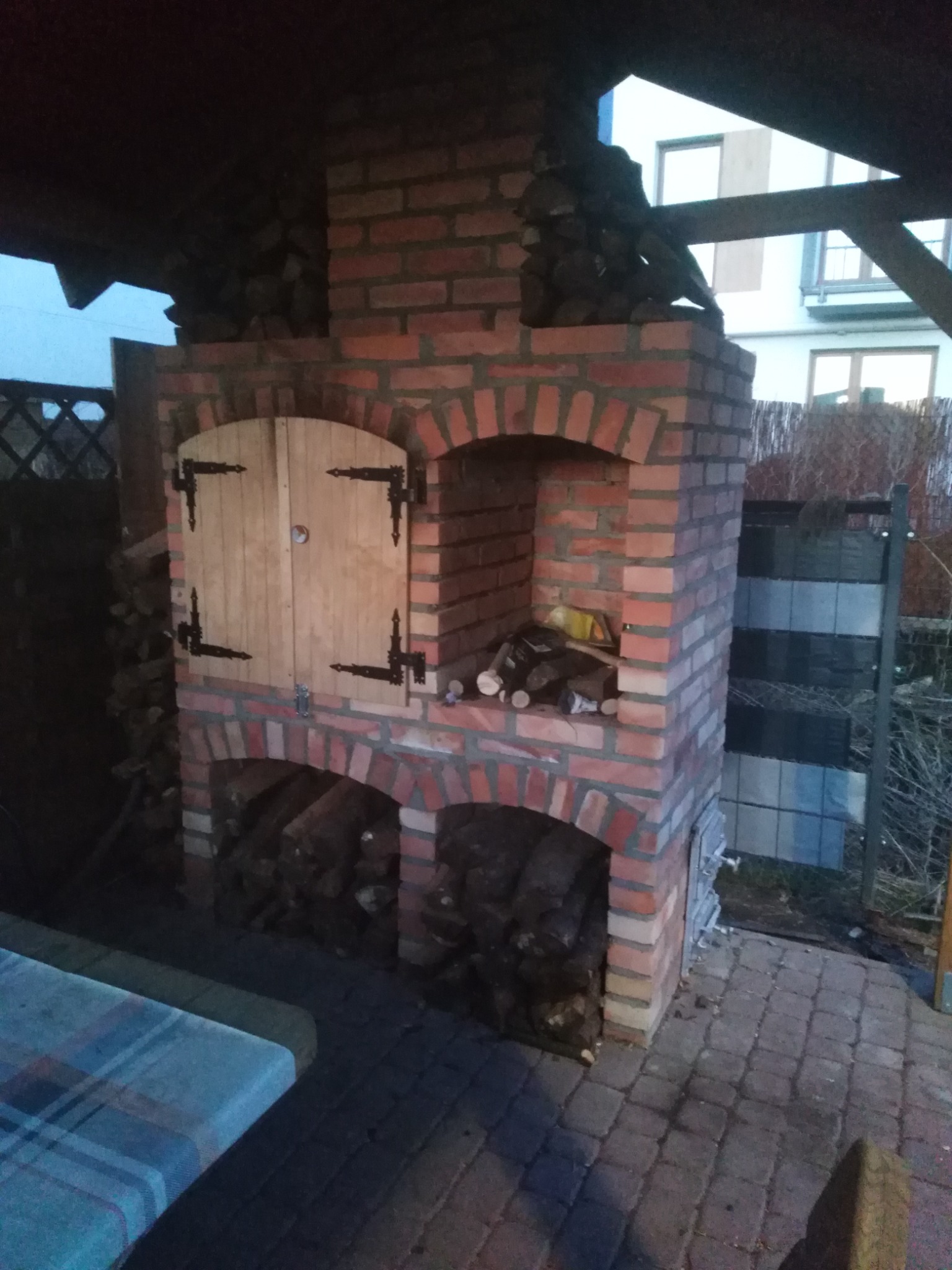 Murowany piec zewnętrzny z czerwonej cegły z drewnianymi drzwiczkami i zapasem drewna, umieszczony na brukowanym patio pod zadaszeniem.