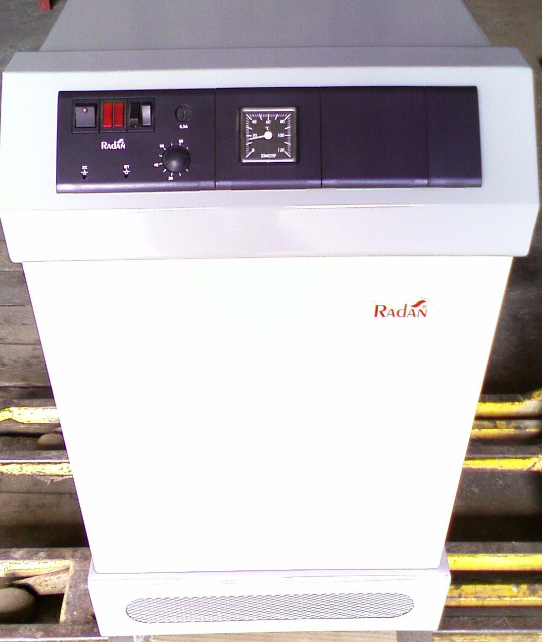 Kocioł gazowy żeliwny Radan RA 22 z widocznym panelem sterowania, manometrem i logotypem producenta na białej obudowie, ustawiony na palecie.