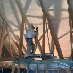 Pracownik w kombinezonie ochronnym aplikuje piankę izolacyjną na poddaszu w Świdnicy, widoczne drewniane krokwie i świeżo nałożona warstwa izolacji.