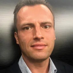 Portret mężczyzny w koszuli w kratę i marynarce na tle ciemnej, teksturowanej ściany. Widoczne zmarszczki mimiczne. Neutralny wyraz twarzy, brązowe oczy.