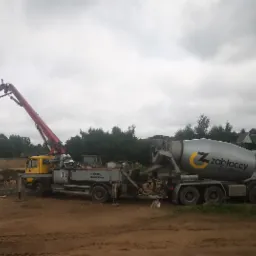 Dwa pojazdy betoniarki: żółta pompa do betonu z wysięgnikiem i niebieska betonomieszarka 'Zabłoccy Beton' na placu budowy, w tle widoczne drzewa i budynki gospodarcze, pochmurne niebo.