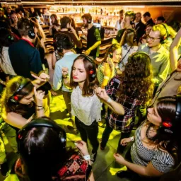 Tłum ludzi tańczących na imprezie Silent Disco, wszyscy w słuchawkach z czerwonymi diodami, bar w tle.