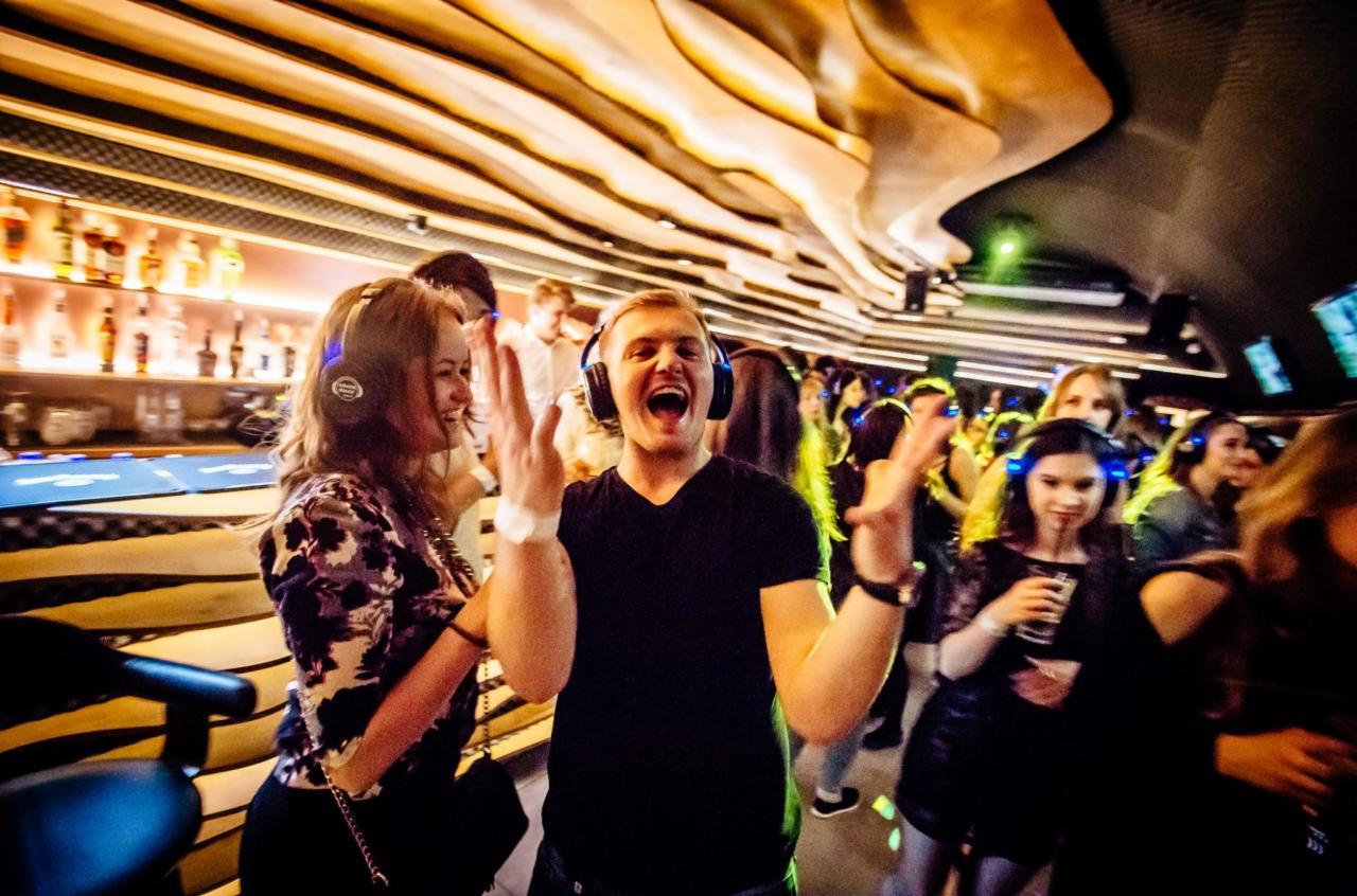 Grupa rozbawionych osób w słuchawkach na imprezie silent disco, bar w tle, dekoracyjne oświetlenie sufitowe.