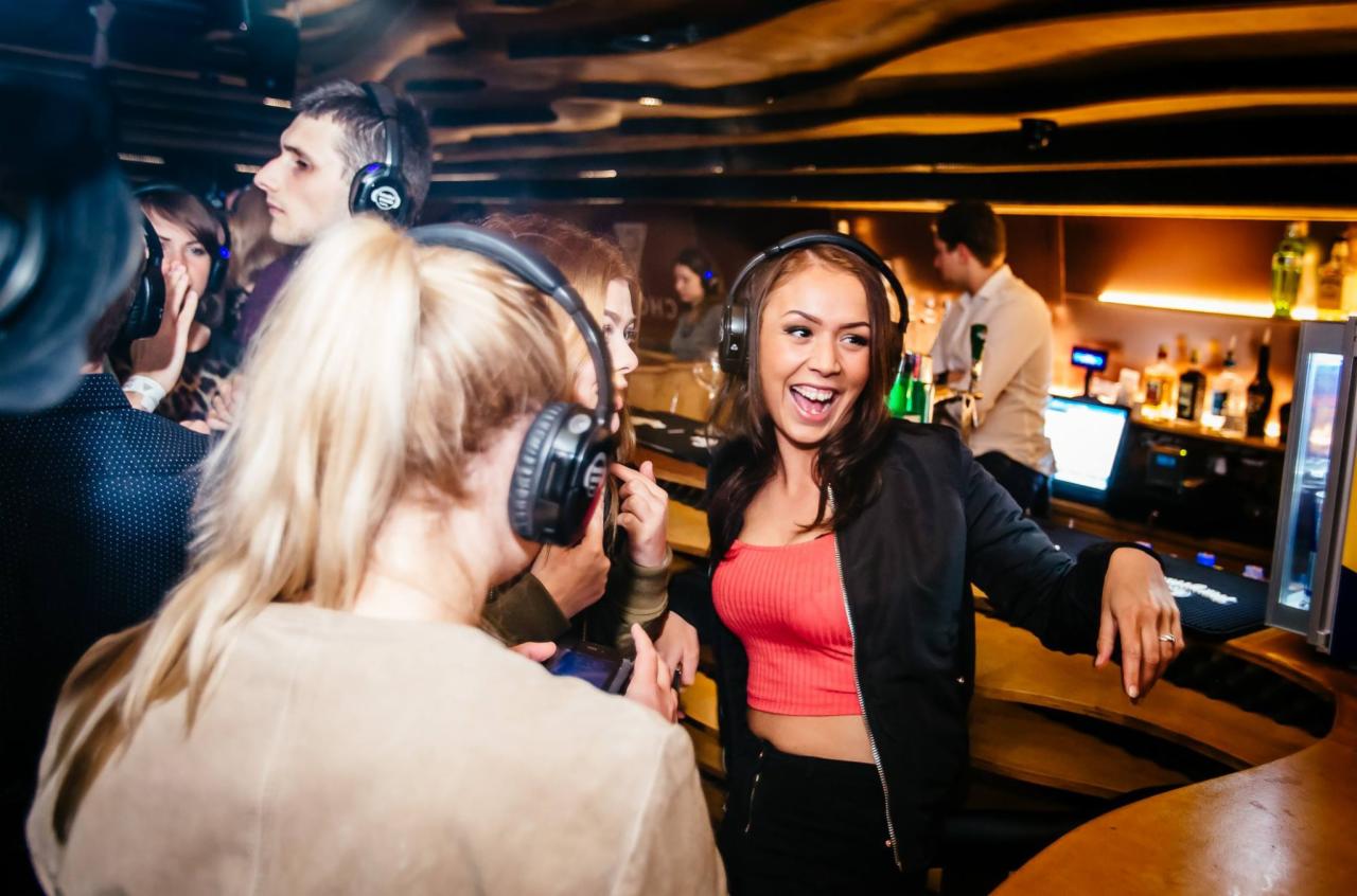 Grupa młodych ludzi w słuchawkach na imprezie typu silent disco, kobieta w czerwonym topie uśmiecha się przy barze.