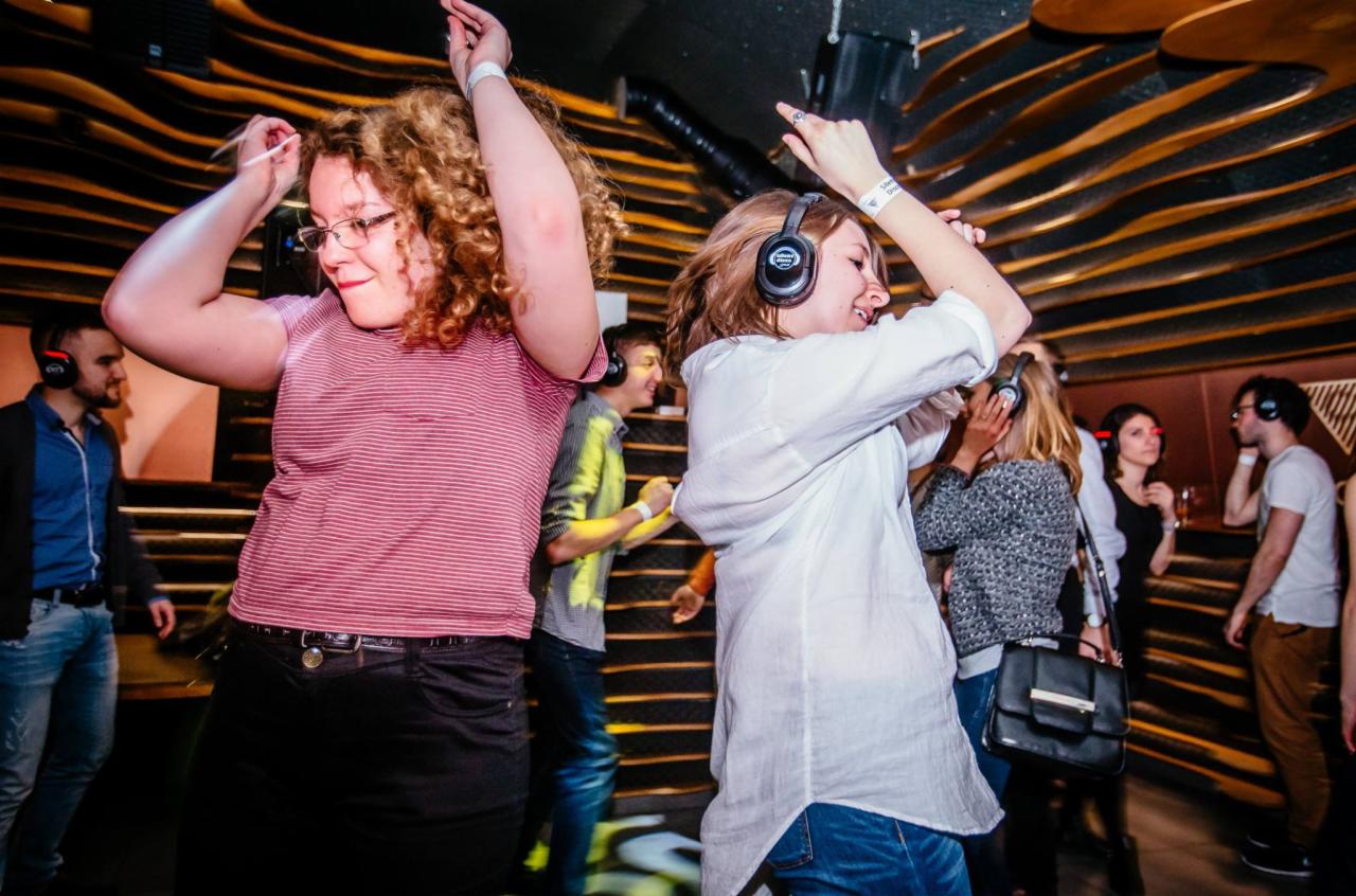 Grupa osób tańczy w słuchawkach na imprezie silent disco w klubie z dekoracyjnym oświetleniem ściennym.