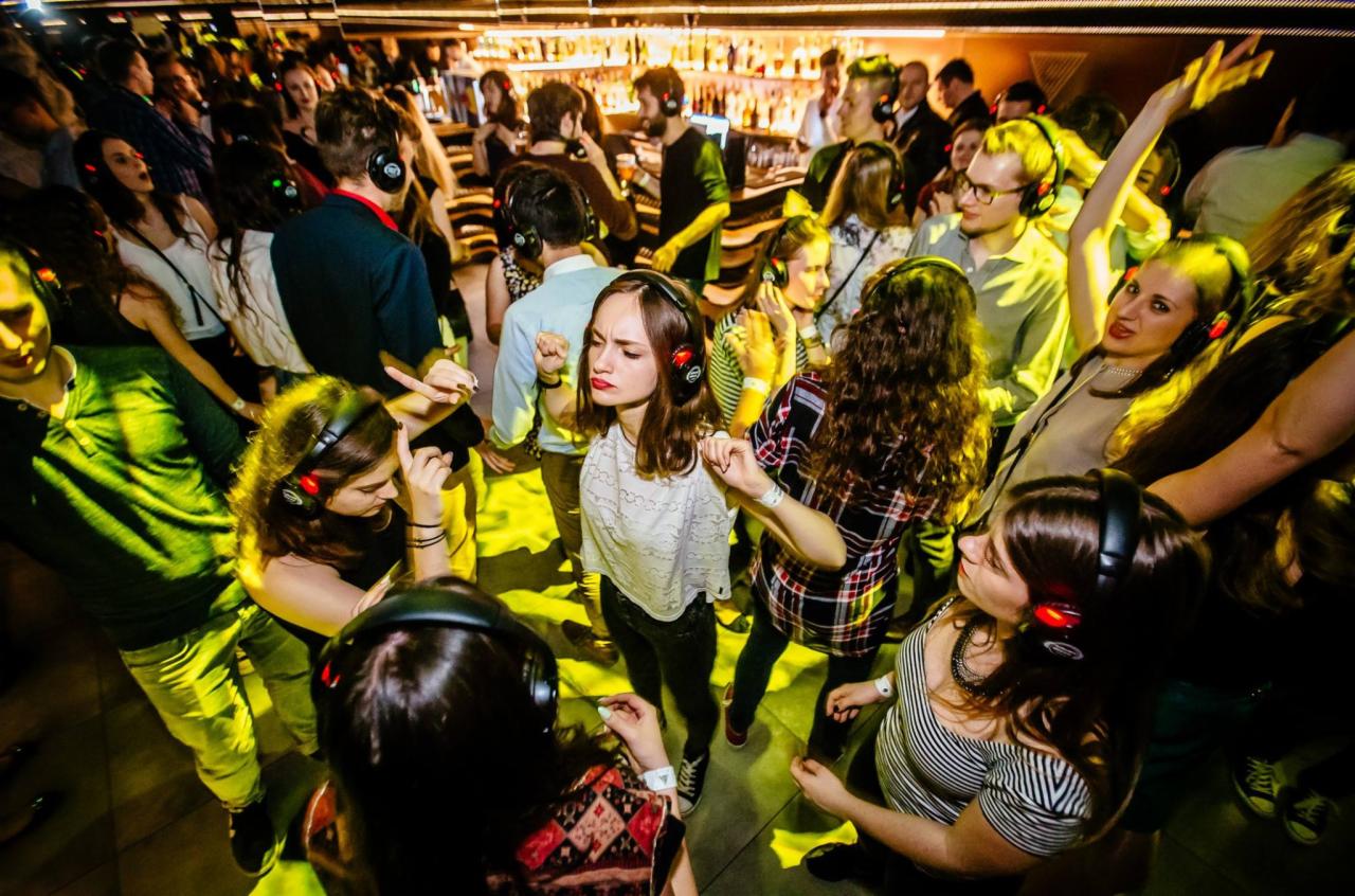 Tłum ludzi tańczących na imprezie Silent Disco, wszyscy w słuchawkach z czerwonymi diodami, bar w tle.