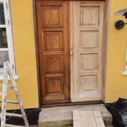 Odnowiona drewniana dwuskrzydłowa brama wejściowa, połowa pomalowana na ciemny brąz, druga połowa w jasnym odcieniu drewna, widoczne schody wejściowe, drabina i żółta elewacja budynku.