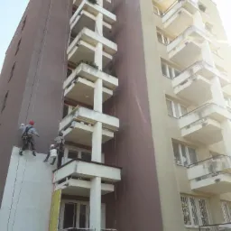 Dwóch alpinistów przemysłowych maluje elewację wysokiego bloku mieszkalnego na brązowo, z widocznymi linami i sprzętem zabezpieczającym.