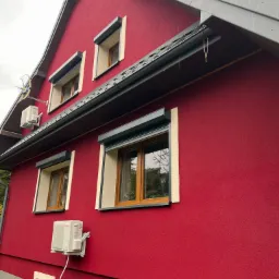 Czerwony dom z oknami z roletami zewnętrznymi, widoczne dwa klimatyzatory na elewacji i szara podmurówka.