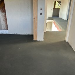 DARBUD - Wnętrze w trakcie budowy: wylana posadzka, otynkowane ściany, widoczne instalacje elektryczne. Surowy stan wykończenia, perspektywa wnętrza z widokiem na schody.