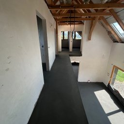 DARBUD - Wnętrze domu w trakcie budowy: świeżo wylana, ciemna posadzka, otynkowane ściany, drewniana konstrukcja dachu z oknami połaciowymi. Widok z góry na otwartą przestrzeń.