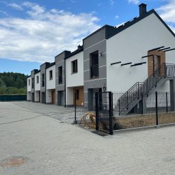 DARBUD - Szeregówka domów z elewacją w odcieniach szarości i bieli, z zewnętrznymi schodami do jednego z lokali. Brukowana droga dojazdowa, błękitne niebo.