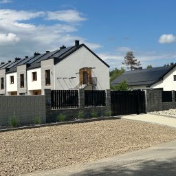 DARBUD - Nowoczesne domy szeregowe z panelami słonecznymi, czarnym ogrodzeniem i kamiennym podłożem. Jeden dom w trakcie budowy, widoczne skrzynki elektryczne.