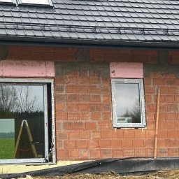 DARBUD - Zewnętrzna ściana domu w trakcie budowy z zamontowanymi oknami z jasnymi ramami, różową izolacją wokół otworów i widoczną czerwoną cegłą. Na górze ciemny dach z oknami połaciowymi.