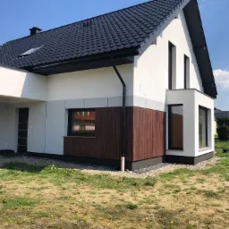 Nowoczesny dom jednorodzinny z białą elewacją, elementami drewnopodobnymi i ciemną dachówką, widok z zewnątrz w słoneczny dzień.