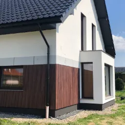 Nowoczesny dom z elewacją w kolorze białym i brązowym drewnem, czarną dachówką i rynnami, widok z boku na tle zieleni i błękitnego nieba.