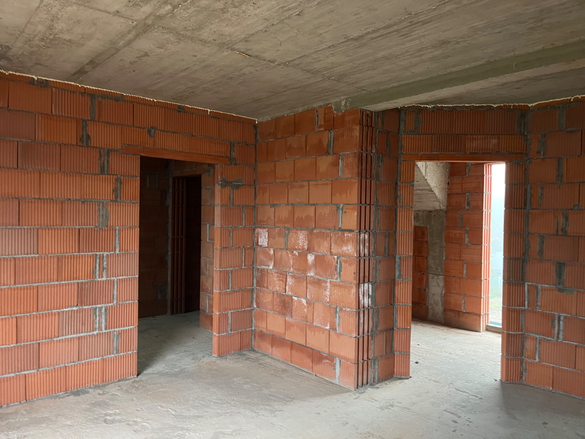Wnętrze surowego domu w trakcie budowy z widocznymi ścianami z czerwonej cegły i betonowym sufitem, przygotowane otwory na drzwi.