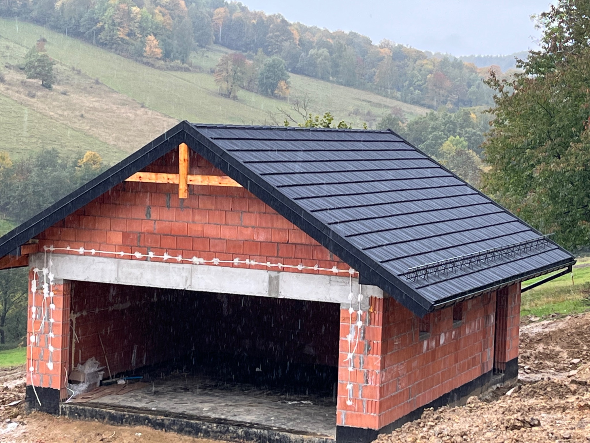 Niedokończony budynek z cegły z czarnym dachem, widoczne elementy konstrukcyjne i instalacje elektryczne, w tle zalesione wzgórze w pochmurny dzień.
