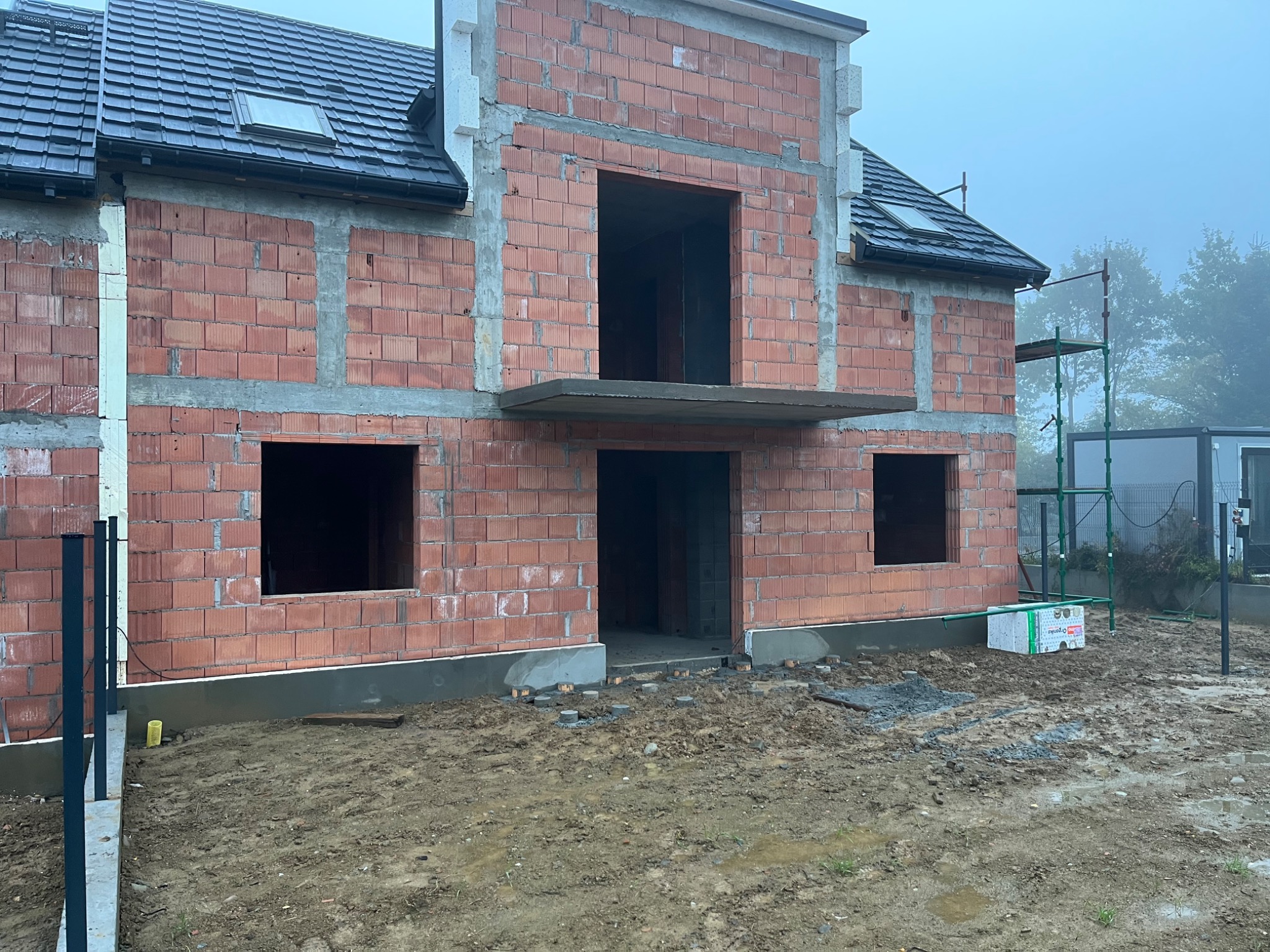 Surowa bryła dwukondygnacyjnego domu w trakcie budowy, ściany z czerwonej cegły ceramicznej, widoczne otwory okienne i drzwiowe, szare fundamenty, częściowo ocieplone, w tle rusztowanie i zarośla...