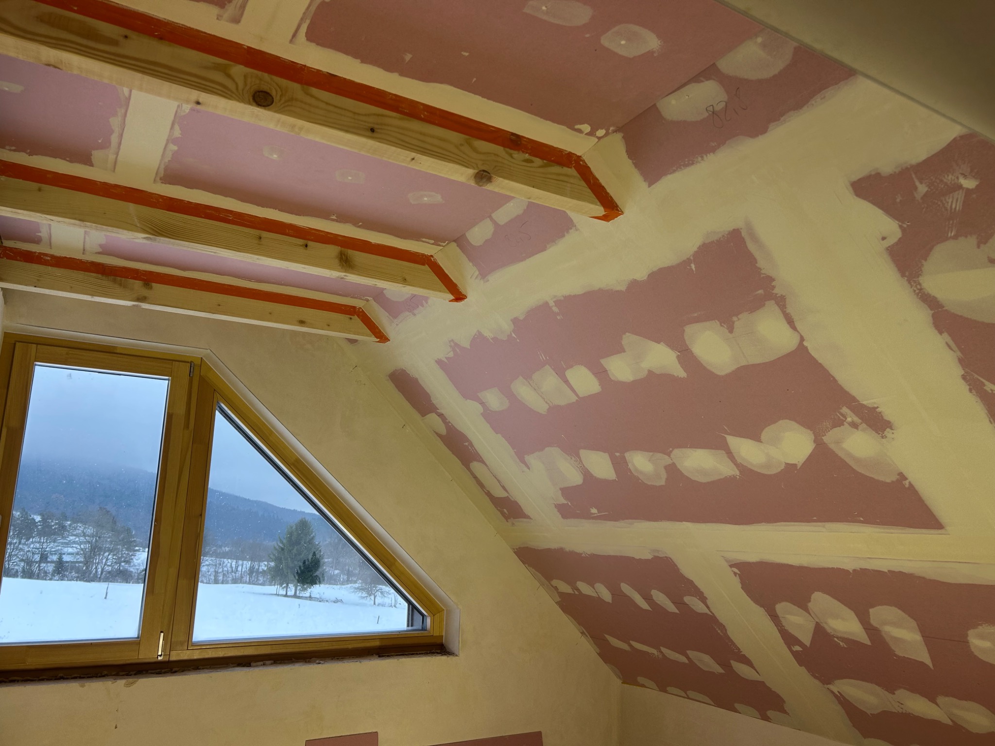 Poddasze w trakcie wykańczania: widoczne płyty gipsowo-kartonowe, drewniane belki stropowe z taśmą, szpachlowane łączenia i okno dachowe z widokiem na zimowy krajobraz.