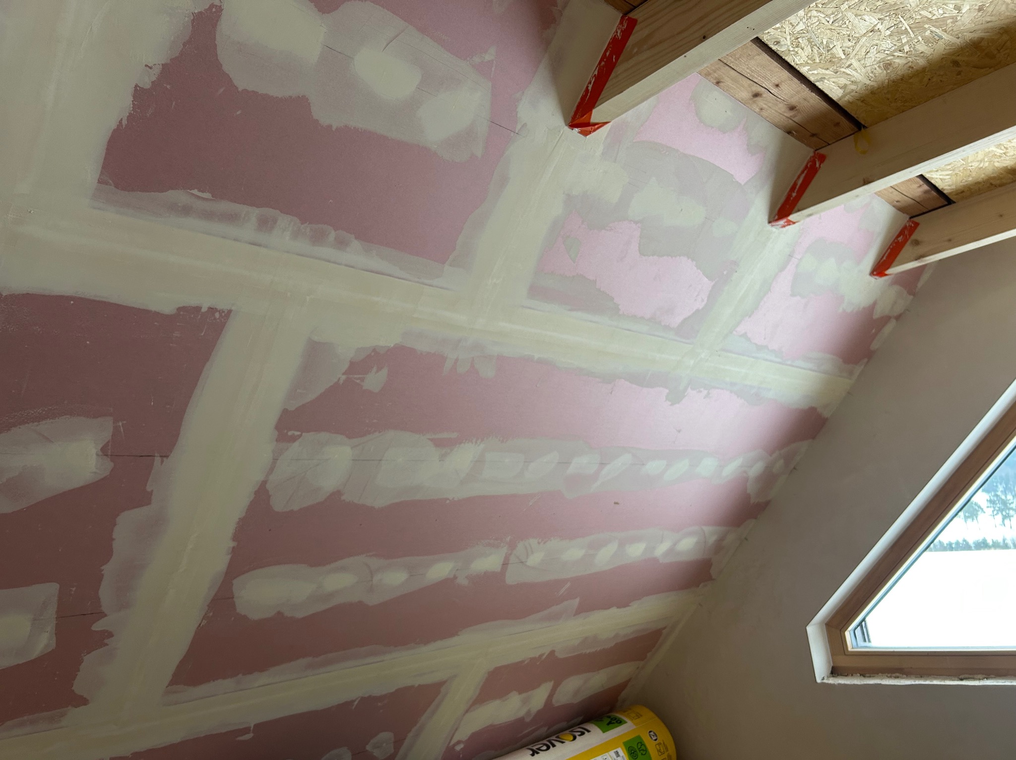 Poddasze z oknem połaciowym w trakcie wykańczania: płyty gipsowe z widocznymi łączeniami, drewniana konstrukcja dachu i rolka izolacji. Prace wykończeniowe.