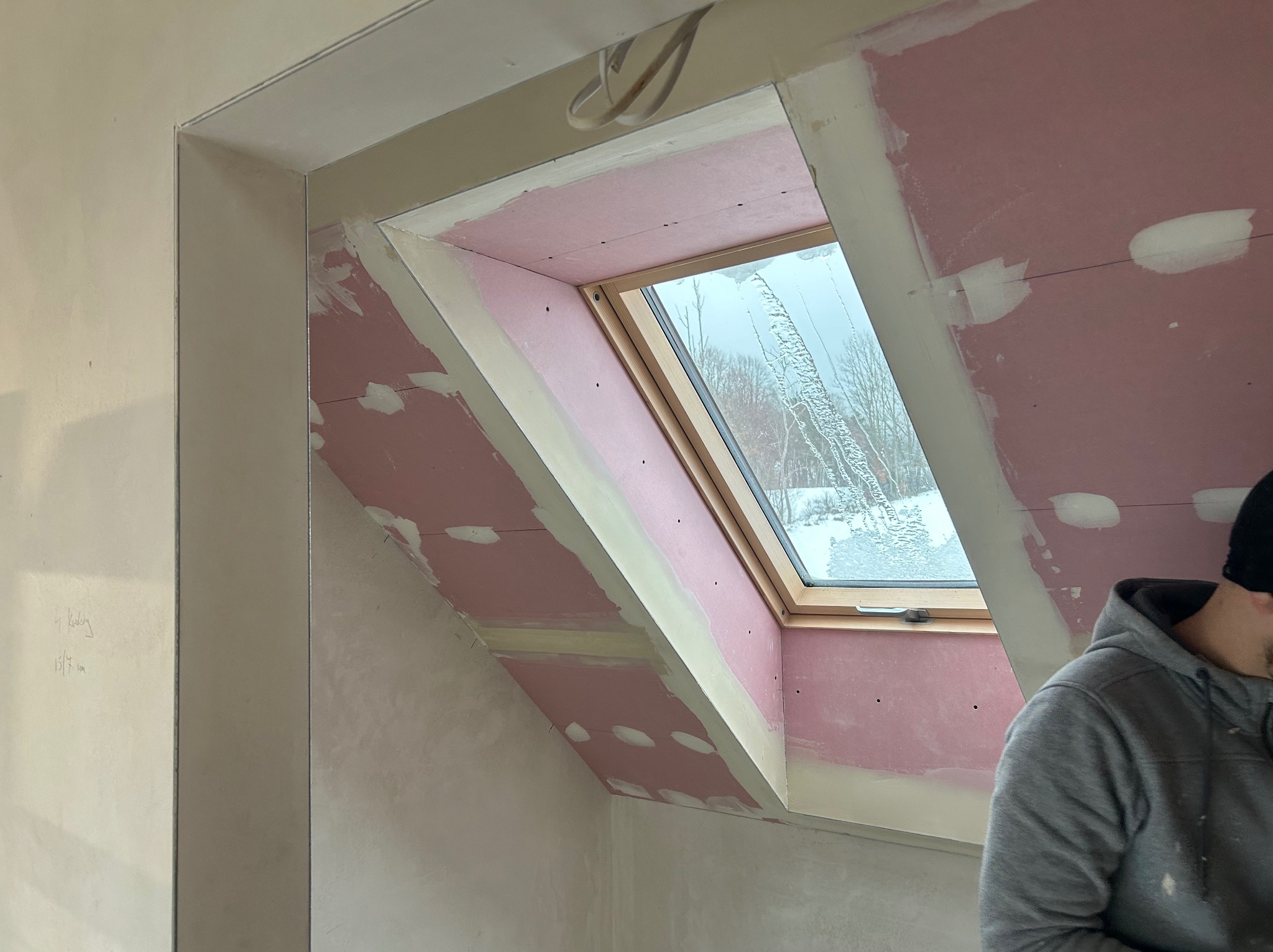 Poddasze z oknem dachowym w trakcie wykańczania: różowe płyty gipsowe, szpachlowanie, widok na zimowy krajobraz. Fragment sylwetki osoby w szarej bluzie.