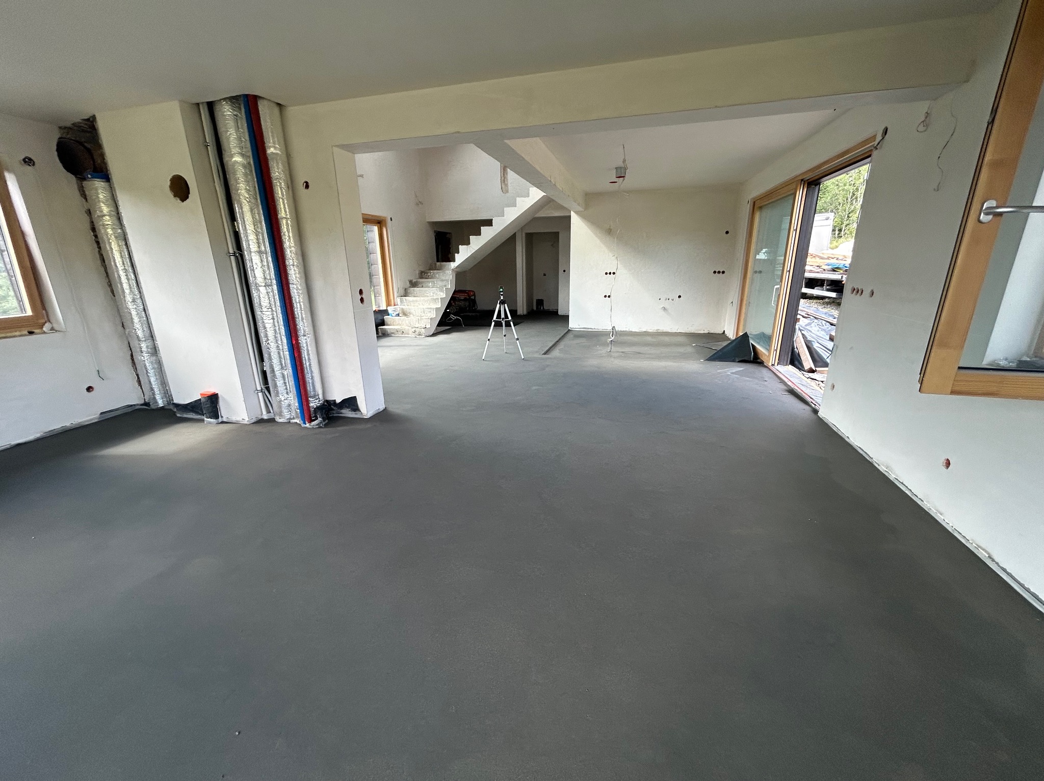 Wnętrze w trakcie prac wykończeniowych: wylewka podłogowa, instalacje przy ścianach, schody w tle. Widoczny laser krzyżowy. Surowy stan, przygotowanie do dalszych prac.