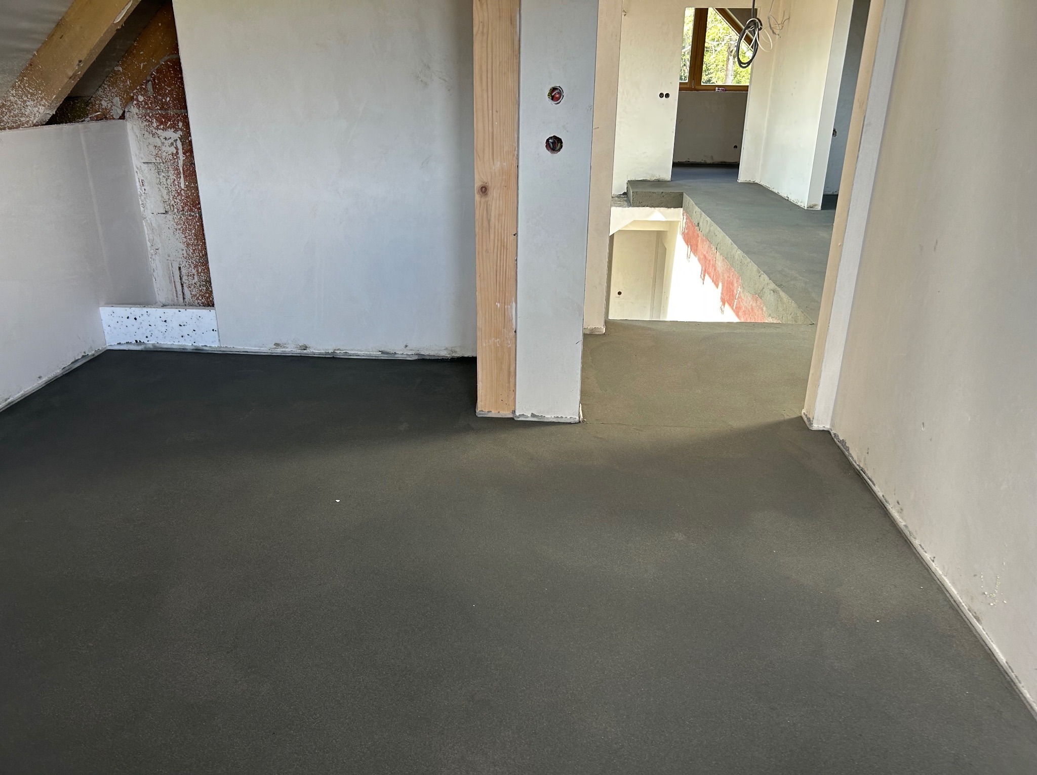 Wnętrze w trakcie budowy: wylana posadzka, otynkowane ściany, widoczne instalacje elektryczne. Surowy stan wykończenia, perspektywa wnętrza z widokiem na schody.