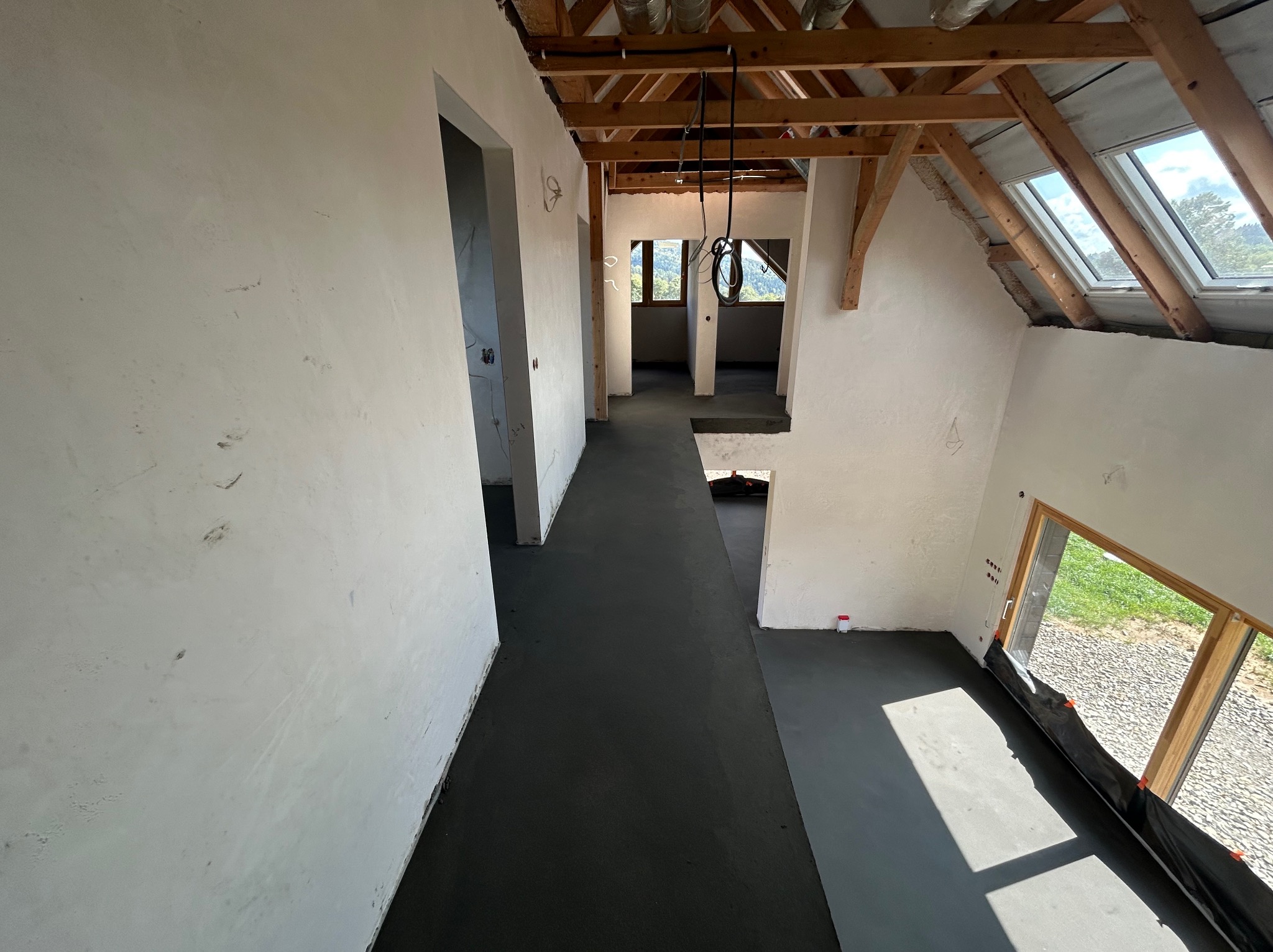 Wnętrze domu w trakcie budowy: świeżo wylana, ciemna posadzka, otynkowane ściany, drewniana konstrukcja dachu z oknami połaciowymi. Widok z góry na otwartą przestrzeń.
