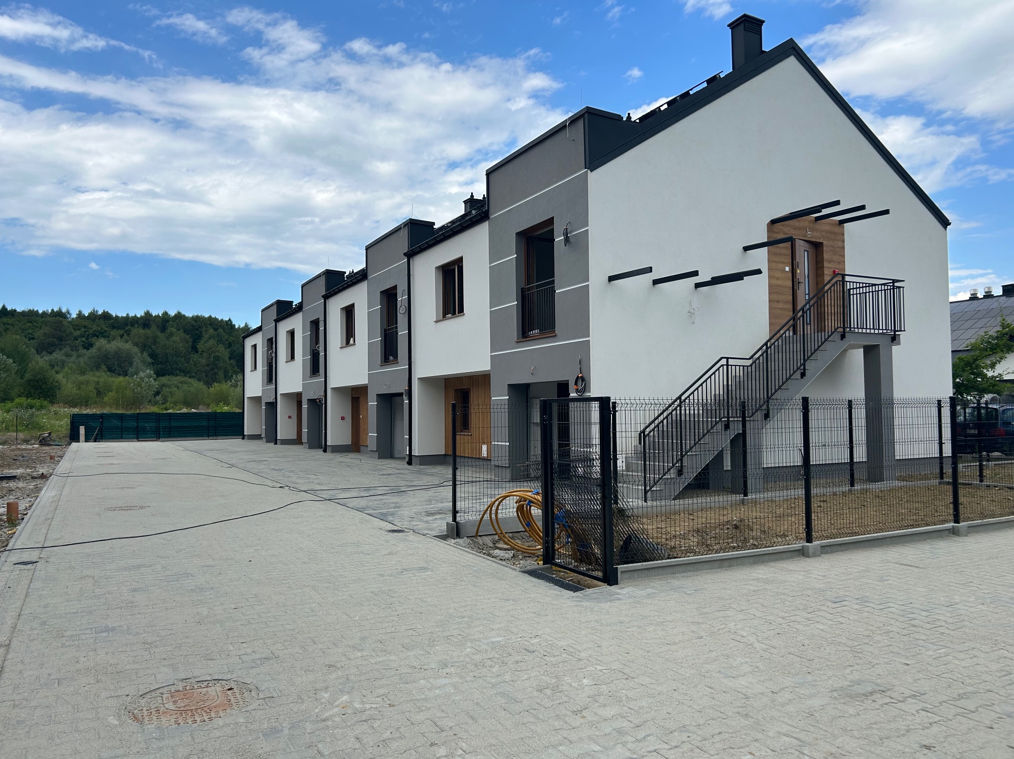 Szeregówka domów z elewacją w odcieniach szarości i bieli, z zewnętrznymi schodami do jednego z lokali. Brukowana droga dojazdowa, błękitne niebo.