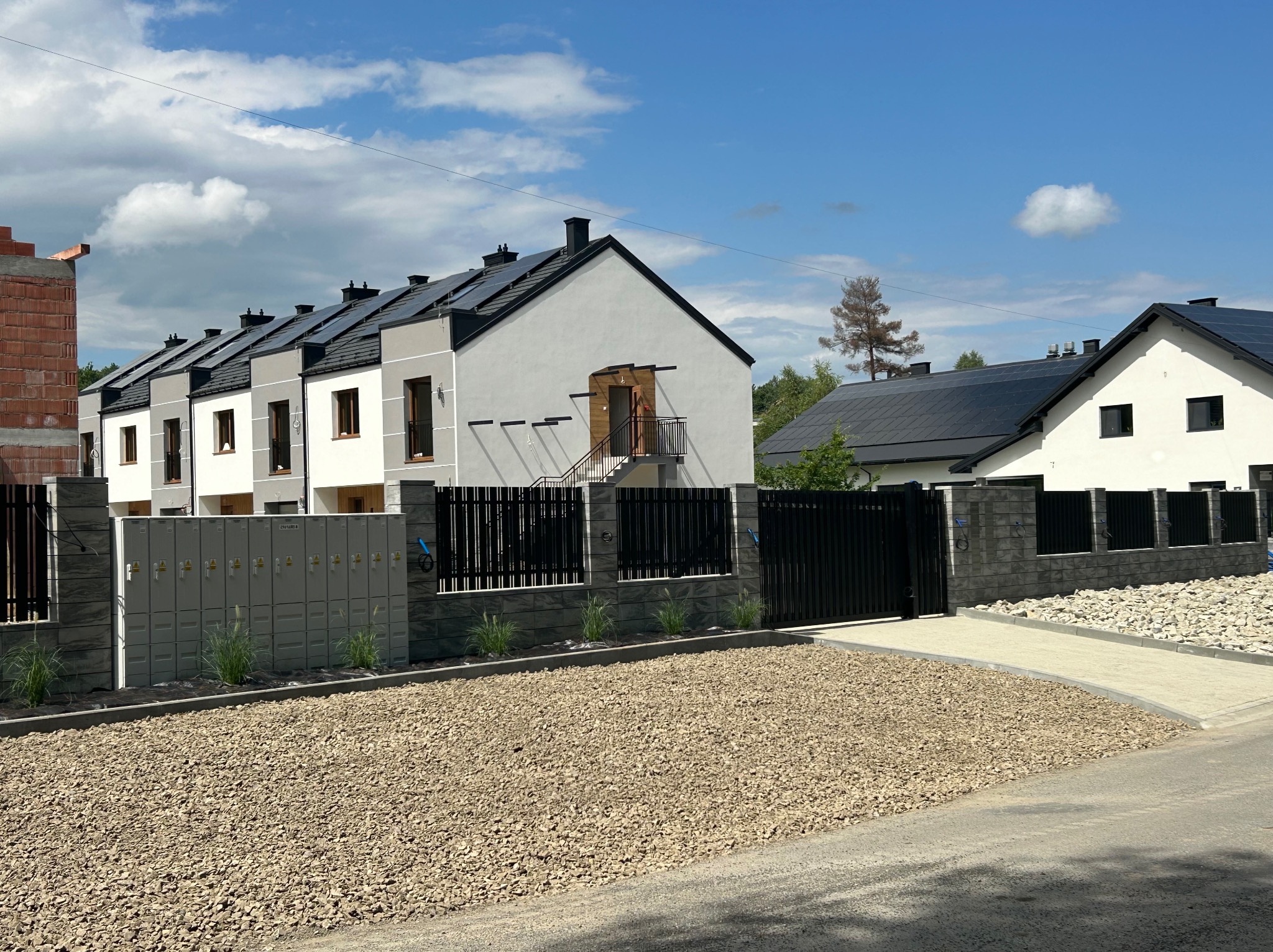 Nowoczesne domy szeregowe z panelami słonecznymi, czarnym ogrodzeniem i kamiennym podłożem. Jeden dom w trakcie budowy, widoczne skrzynki elektryczne.