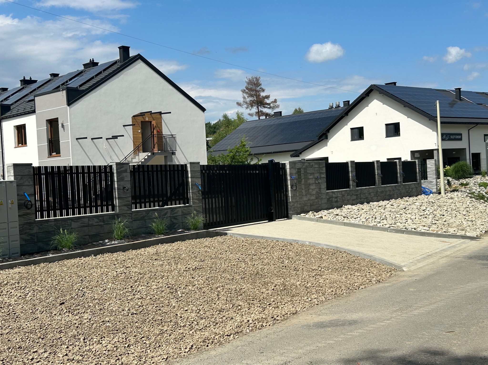 Nowoczesne domy jednorodzinne z panelami słonecznymi, ogrodzone czarnym płotem z kamiennymi słupkami. Podjazd wyłożony jasnym kamieniem.
