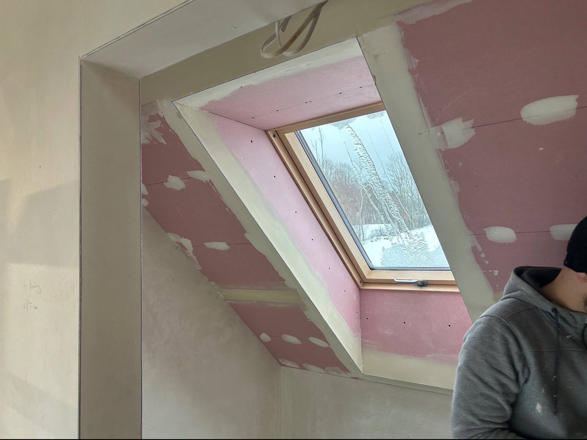 Poddasze w trakcie wykańczania: zamontowane okno dachowe z jasną drewnianą ramą, otoczone różowymi płytami gipsowo-kartonowymi z widocznymi śladami szpachlowania. Fragment osoby w szarej bluzie.