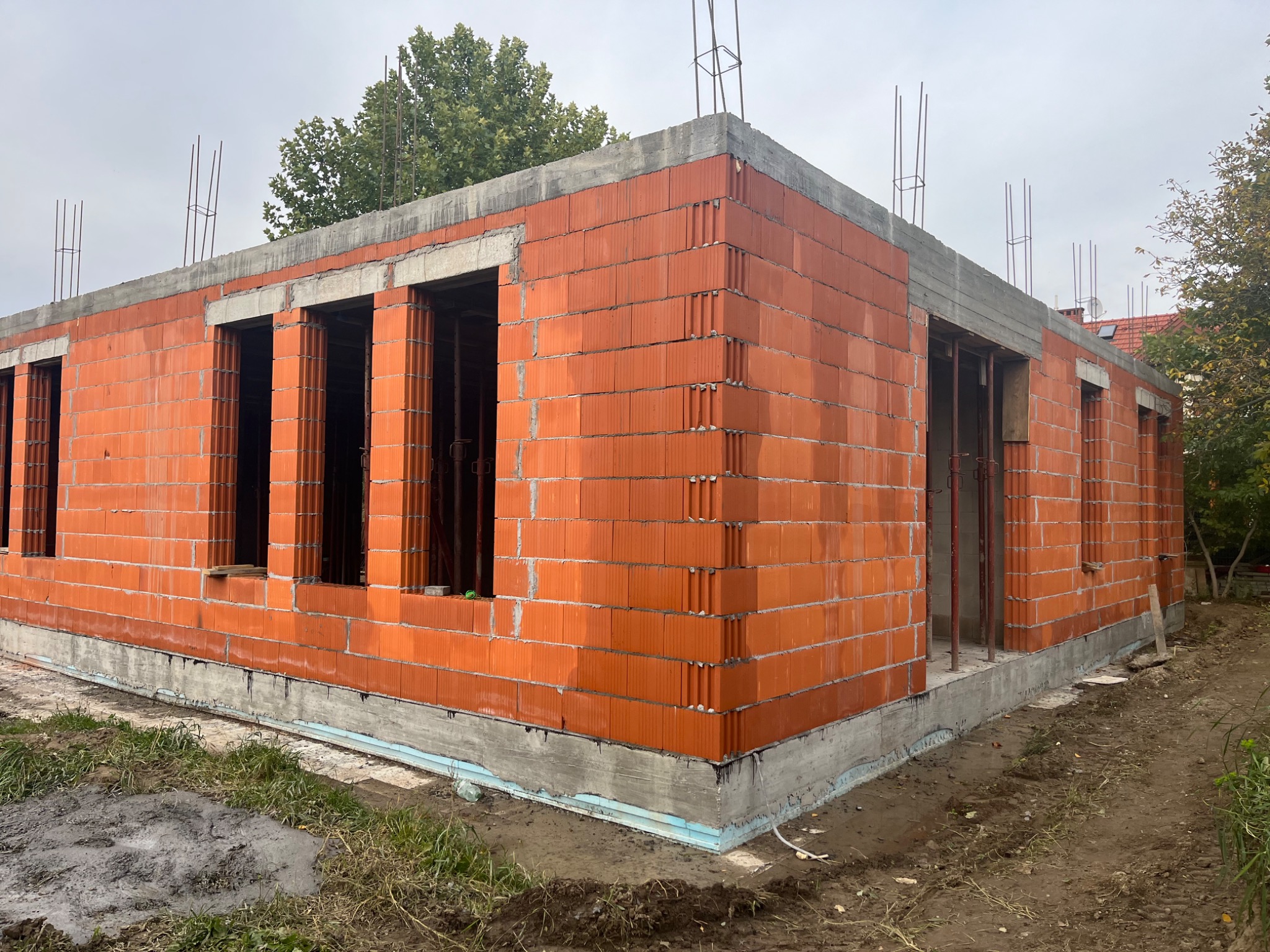 Surowy stan domu z czerwonej cegły ceramicznej, widoczne otwory okienne i pręty zbrojeniowe wystające z betonowego stropu, fundament ocieplony styropianem, w tle drzewa i pochmurne niebo.