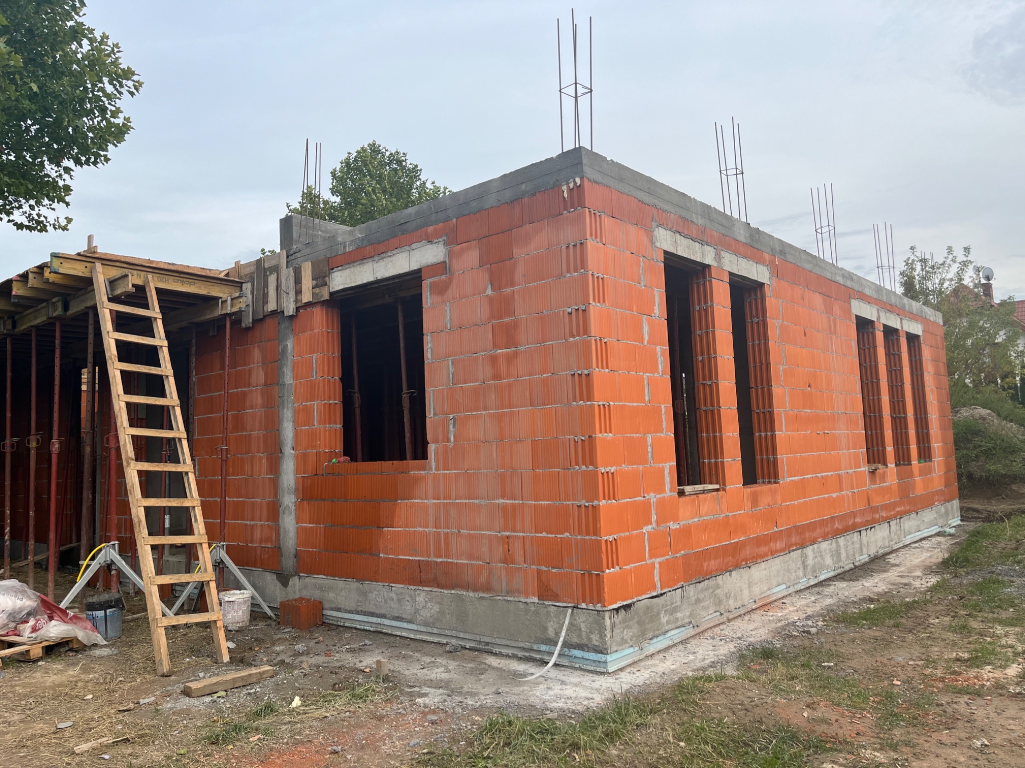 Surowy budynek z czerwonej cegły ceramicznej w trakcie budowy, widoczne otwory okienne, wystające pręty zbrojeniowe na górze, drewniana drabina i szalunek z lewej strony, teren wokół budynku...