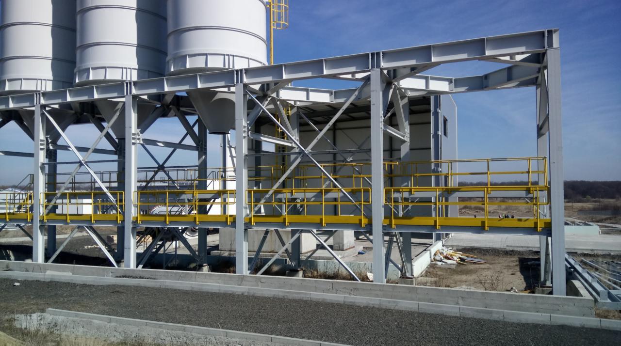 Konstrukcja stalowa z żółtymi barierkami na tle silosów i błękitnego nieba.