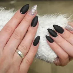 Dłonie z czarnymi, matowymi paznokciami w kształcie migdała, jeden paznokieć z brokatem, srebrny pierścionek na palcu, na tle białej, puszystej tkaniny.