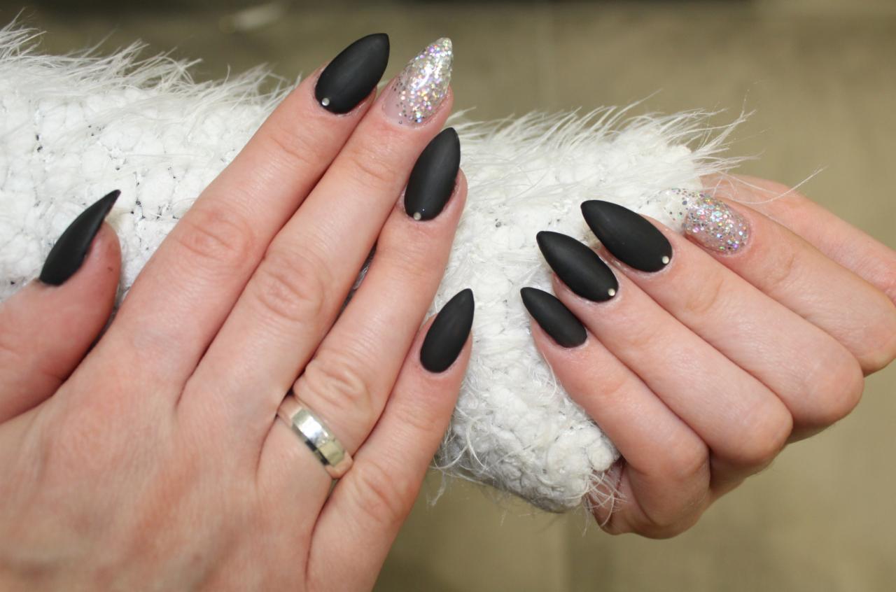 Dłonie z czarnymi, matowymi paznokciami w kształcie migdała, jeden paznokieć z brokatem, srebrny pierścionek na palcu, na tle białej, puszystej tkaniny.