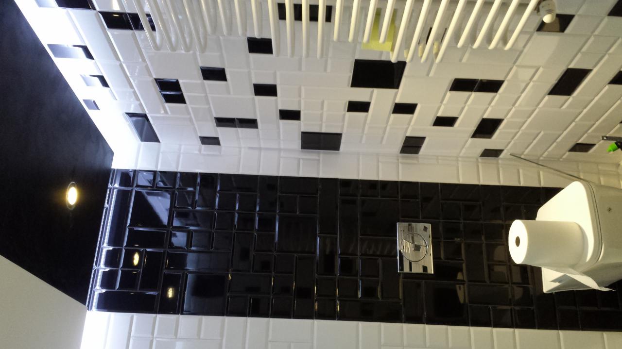 Czarno-biała łazienka z błyszczącymi, czarnymi płytkami na ścianie i białymi płytkami z czarnymi akcentami na suficie, widoczny dozownik papieru toaletowego i panel sterujący spłuczką.