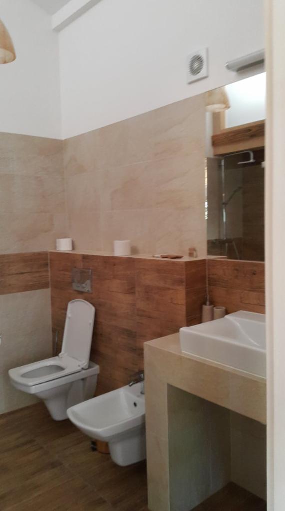 Wykończona łazienka z toaletą, bidetem i umywalką na postumencie. Ściany wyłożone płytkami imitującymi drewno i beżowym kamieniem. Widoczny fragment lustra i wentylator na suficie.