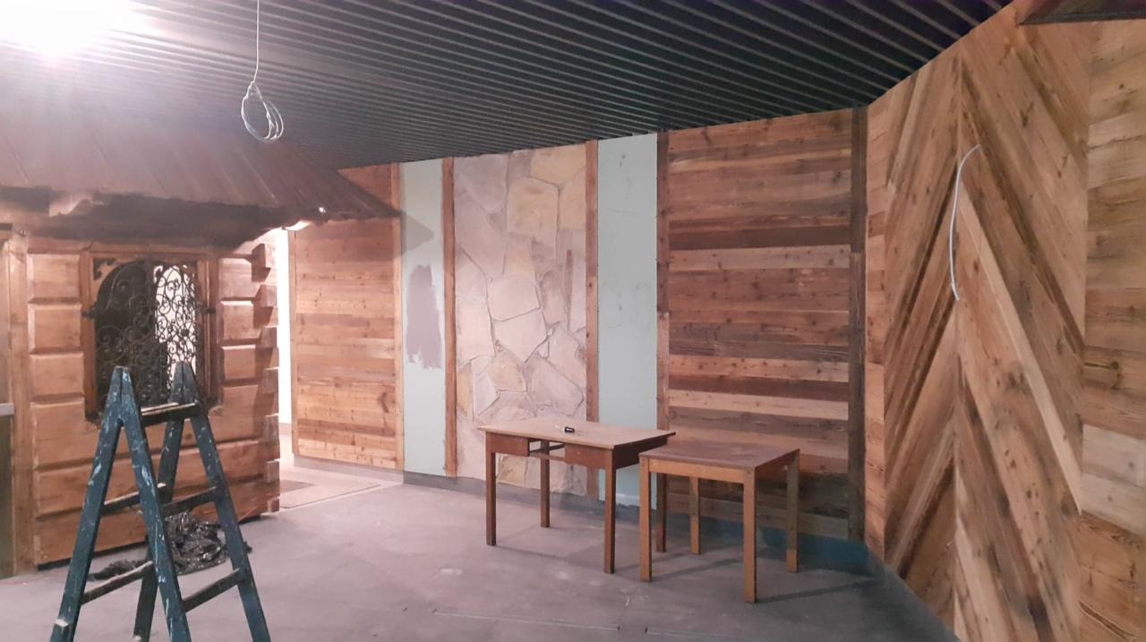 Wnętrze w trakcie remontu: drewniana boazeria w różnych wzorach, fragment ściany wyłożony kamieniem, drewniana konstrukcja przypominająca wejście do domku, drabina malarska i dwa stoliki...