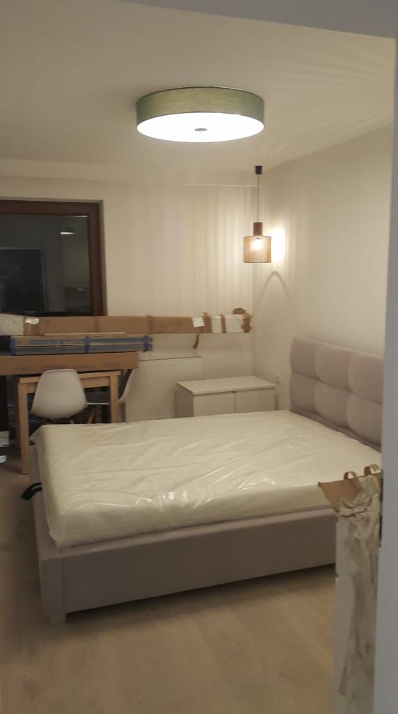 Nowe łóżko z szarym zagłówkiem i materacem w folii ochronnej w pokoju podczas remontu, widoczny stolik nocny, lampa sufitowa i wisząca, okno oraz zabezpieczone kartonami elementy