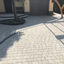 Jasnoszara kostka brukowa ułożona na podjeździe z elementami dekoracyjnymi w postaci ciemnoszarej obwódki i okręgu wokół drzewa, widoczne cienie drewnianej konstrukcji.