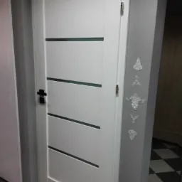 Białe drzwi wewnętrzne z poziomymi przeszkleniami, czarna klamka, zamontowane w szarej ścianie z dekoracyjnymi sztukateriami, widoczna szachownica podłogowa.